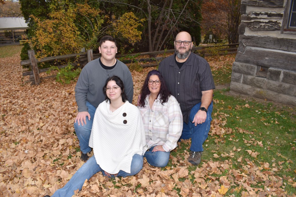 Image is of The Richardson Family, all together.  Darrin and Chris are in the back wearing gray shirts with black t-shirts and blue jeans.  Caroline is wearing a white cowl neck poncho sweater and blue jeans, and Amy Sara is wearing a flannel shirt with muted pink, gray, and purple plaid and blue jeans.  The background is a beautiful fall day in Sunset Park in Washington Iowa by the DAR Cabin.